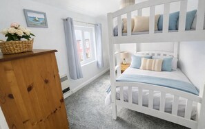 Ein Etagenbett mit Kissen, eine Holzkommode, ein Blumenkorb, ein Fenster mit Vorhängen und ein Gemälde an der Wand.