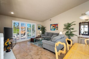 A living room with a sofa, coffee table, TV, and a dining table with chairs. - Wilson's Desert Oasis - Beautiful 3-bdrm condo in Palm Desert with AC, amenities (Palm Desert)