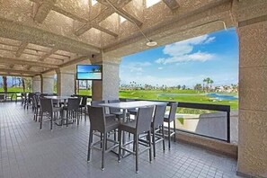 An outdoor patio area with tables and chairs, a TV screen, and a view of a golf course. - Wilson's Desert Oasis - Beautiful 3-bdrm condo in Palm Desert with AC, amenities (Palm Desert)
