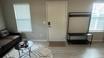 A living room with a ceiling fan, a door, a window with blinds, a brown leather sofa, a coffee table with a decorative item, and a black metal shelf.