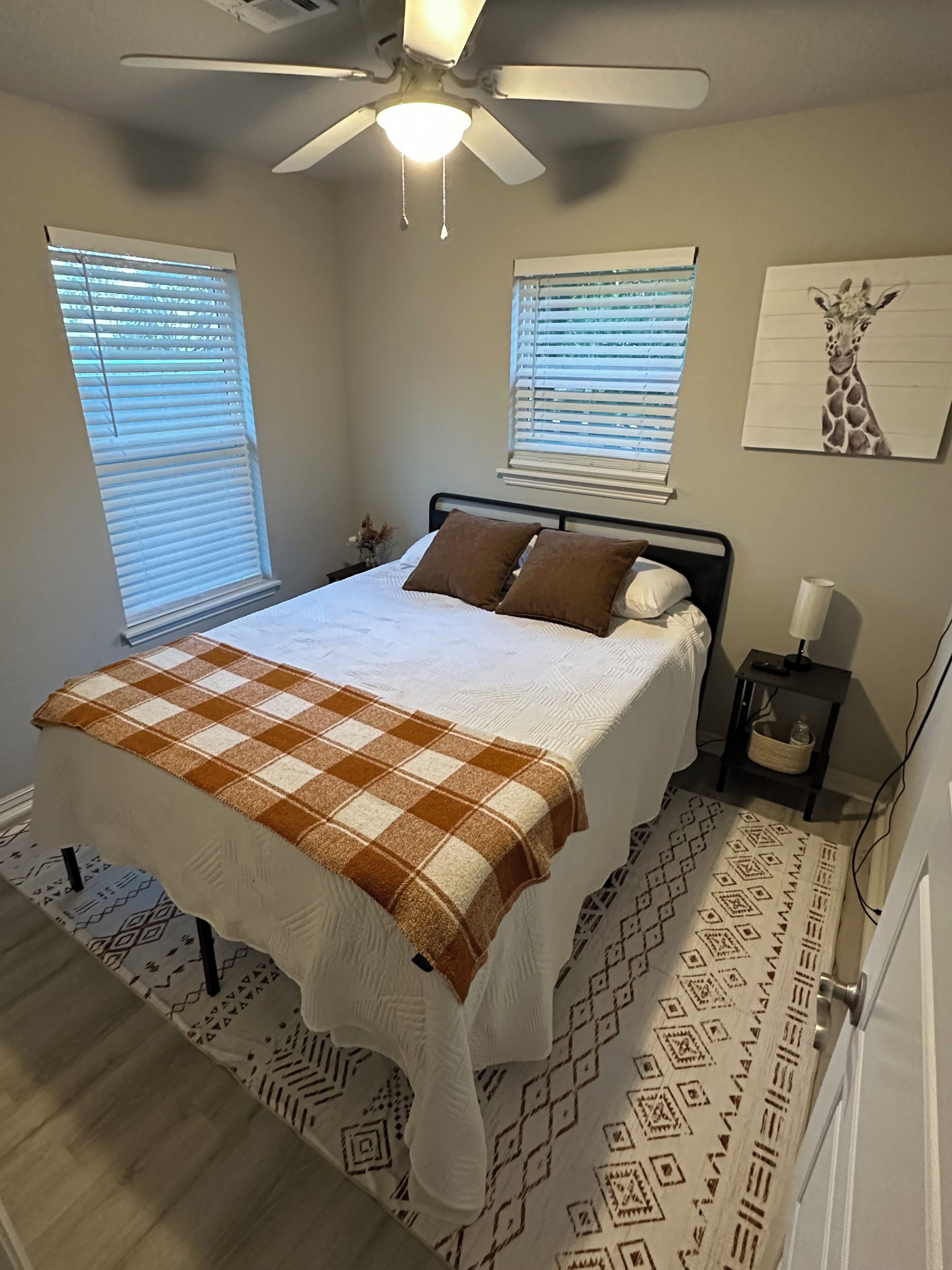 A bedroom with a bed, two windows with blinds, a ceiling fan, a wall-mounted artwork of a giraffe, and a patterned rug.