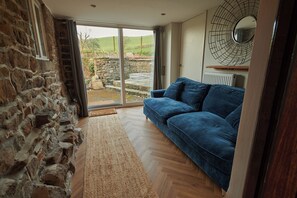 Une pièce avec un mur en pierre, un canapé bleu, un plancher en bois et une vue sur un jardin, visible à travers une porte vitrée.