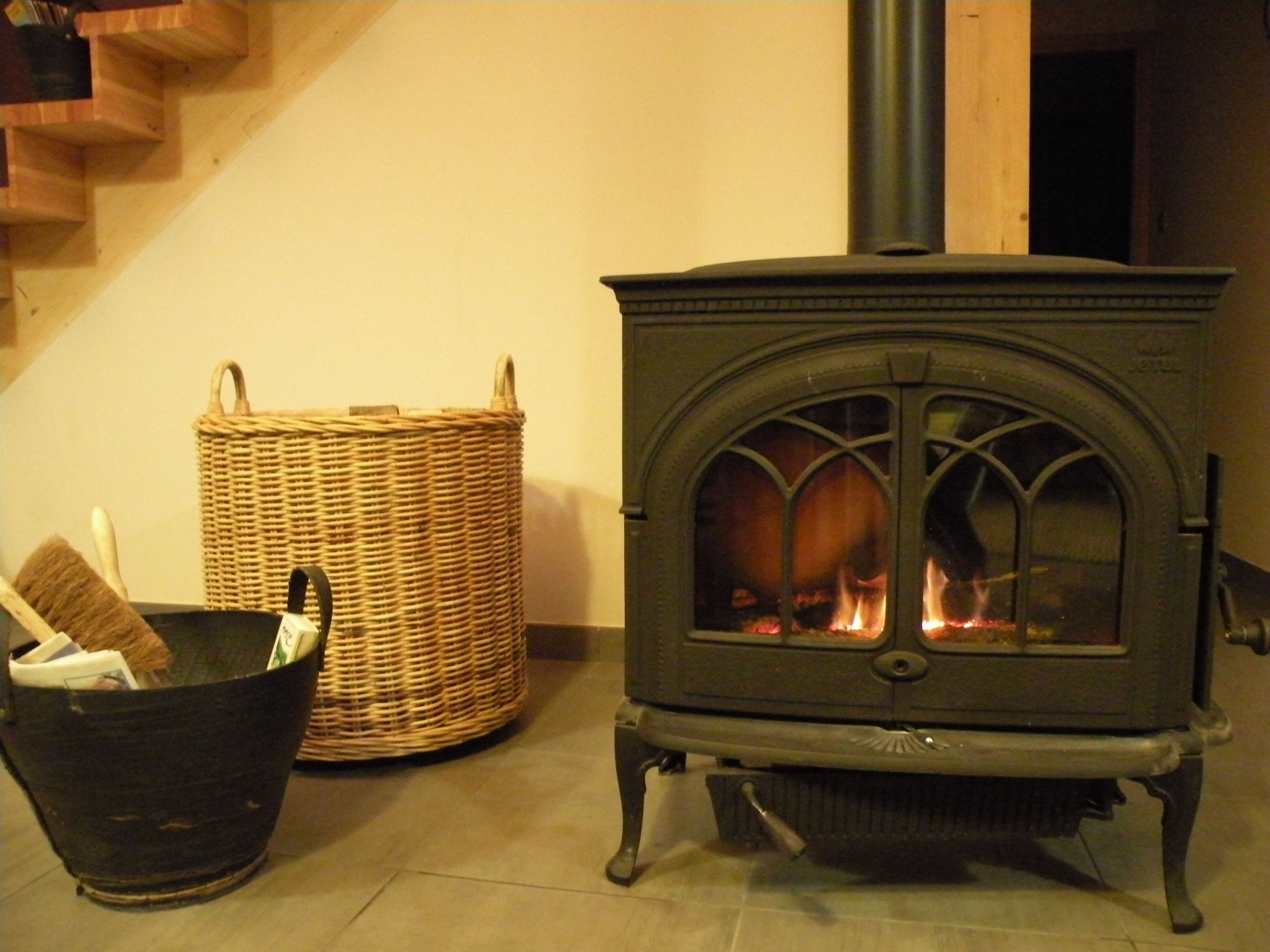 A wood-burning stove with a glass door, a wicker basket, and a black trash bin in a room with a wooden staircase.