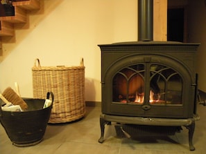 A wood-burning stove with a glass door, a wicker basket, and a black trash bin in a room with a wooden staircase.