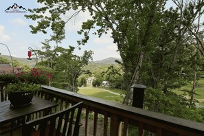 A wooden deck with a view of a grassy area and trees, a red bird feeder, and a building in the distance. - Slopeside & Creekside Sugar Mountain Condo (Sugar Mountain)