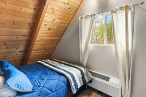 Una habitación pequeña con techo inclinado, una cama, un ventanal con cortinas y un calefactor en la pared.