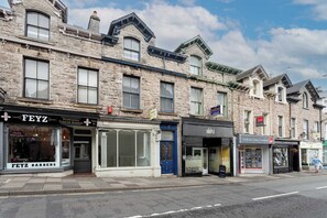 A row of traditional stone buildings with various shops, including a barbershop and a property sales office. - Charming 2BR Flat on Grange-Over-Sands High Street (Grange over Sands)