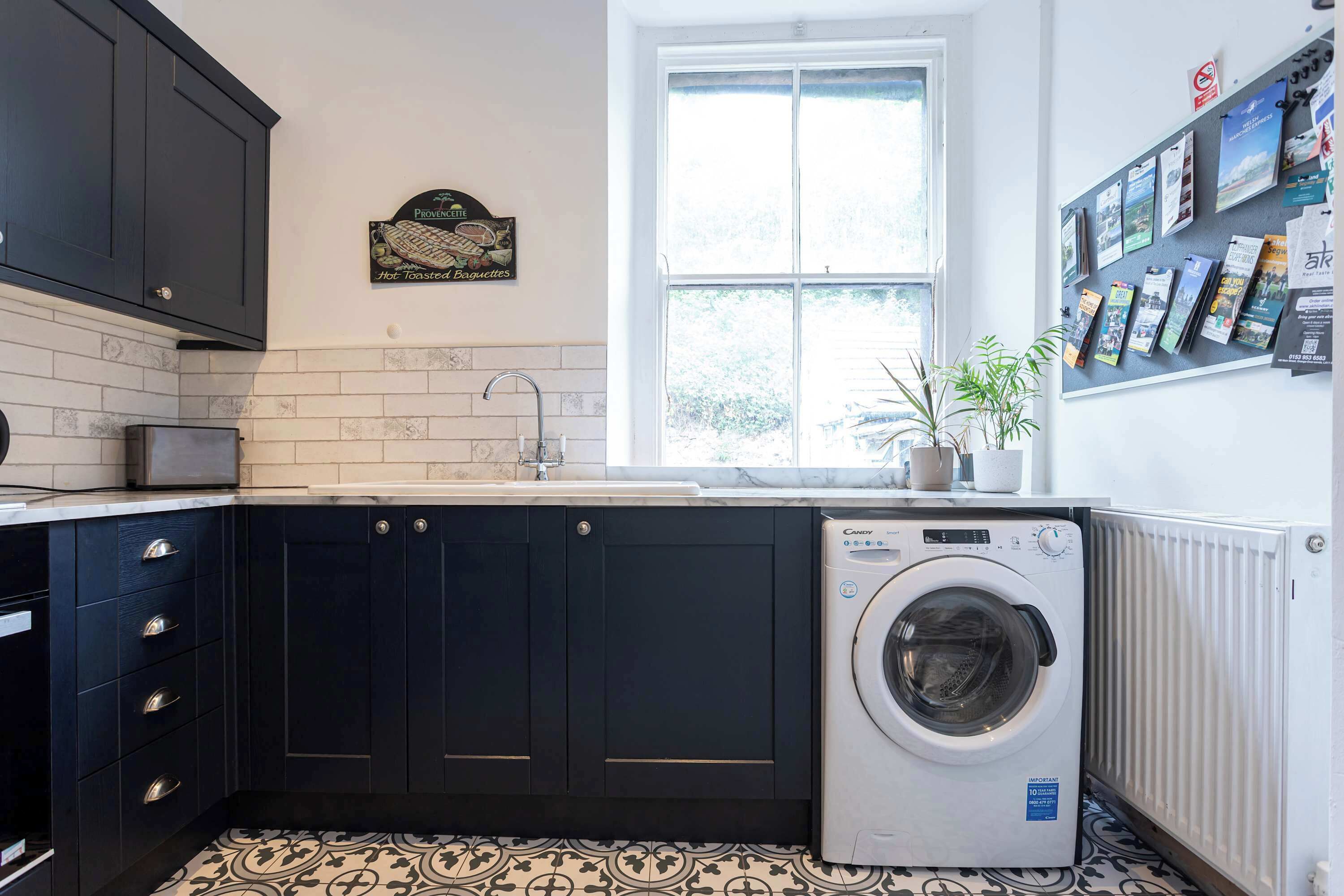 A compact kitchen with dark cabinetry, a built-in washing machine, a window with a view, and a bulletin board with travel brochures.