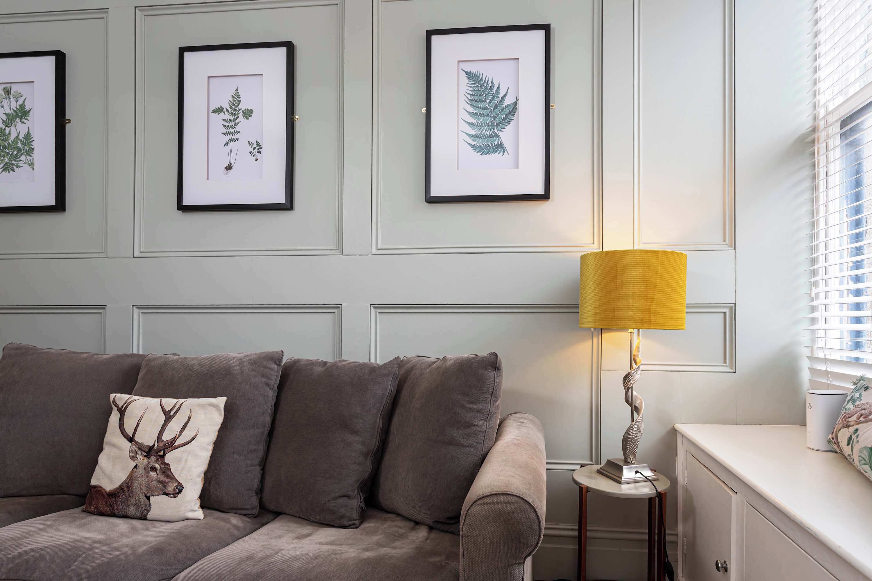 A room with a grey sofa, a lamp, and framed botanical prints on the wall.