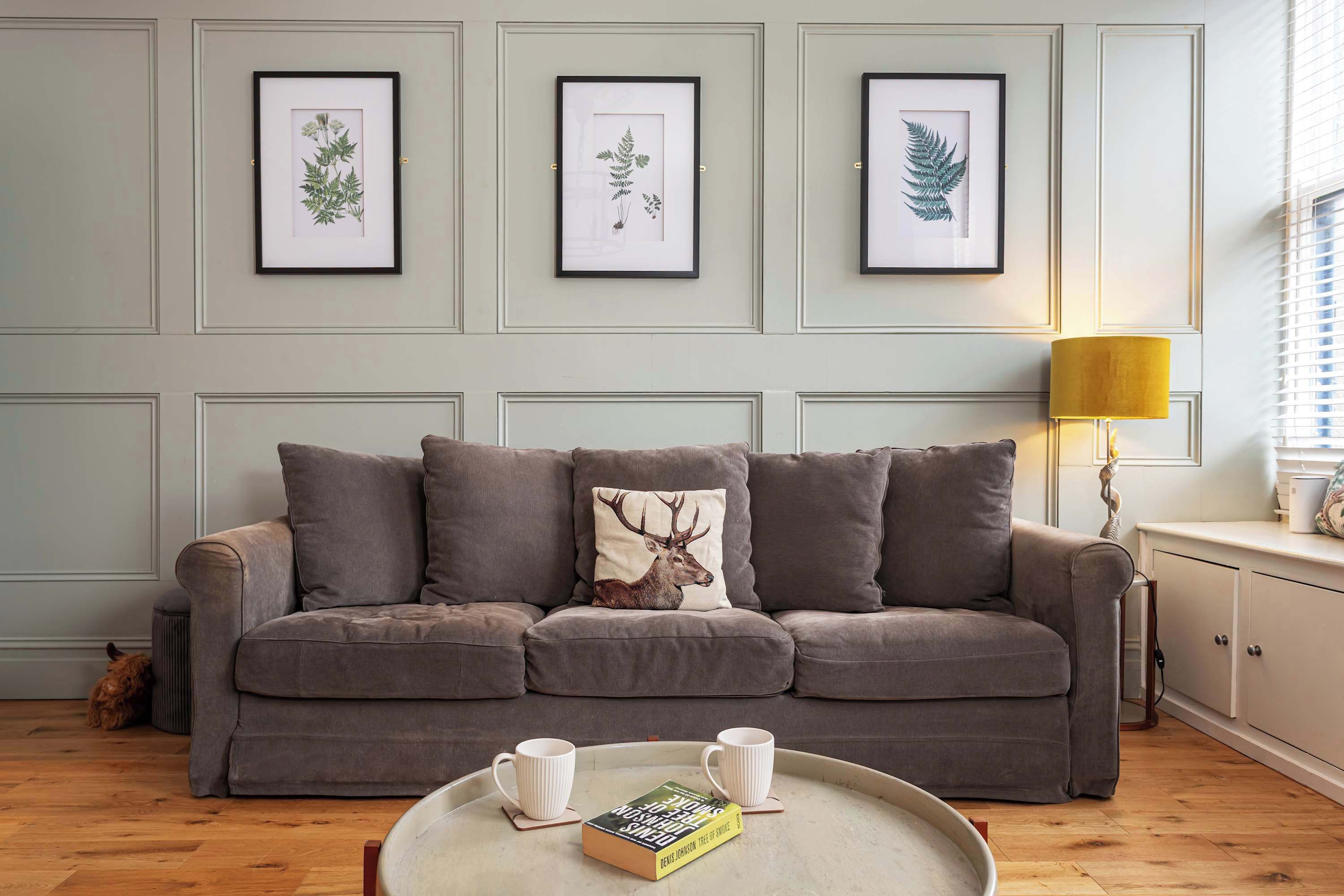 A living room with a grey sofa, framed botanical prints on the wall, a lamp, and a tray with a book and two mugs.
