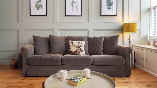 A living room with a grey sofa, framed botanical prints on the wall, a lamp, and a tray with a book and two mugs. - Charming 2BR Flat on Grange-Over-Sands High Street (Grange over Sands)