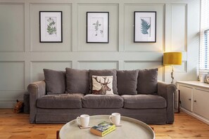 Un salon avec un canapé gris, des reproductions botaniques encadrées accrochées au mur, une lampe et un plateau sur lequel se trouvent un livre et deux tasses.