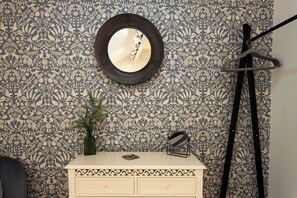 Une salle de bain avec un papier peint à motifs, un miroir rond, un meuble-lavabo blanc avec des tiroirs, une plante verte et un porte-serviettes noir.