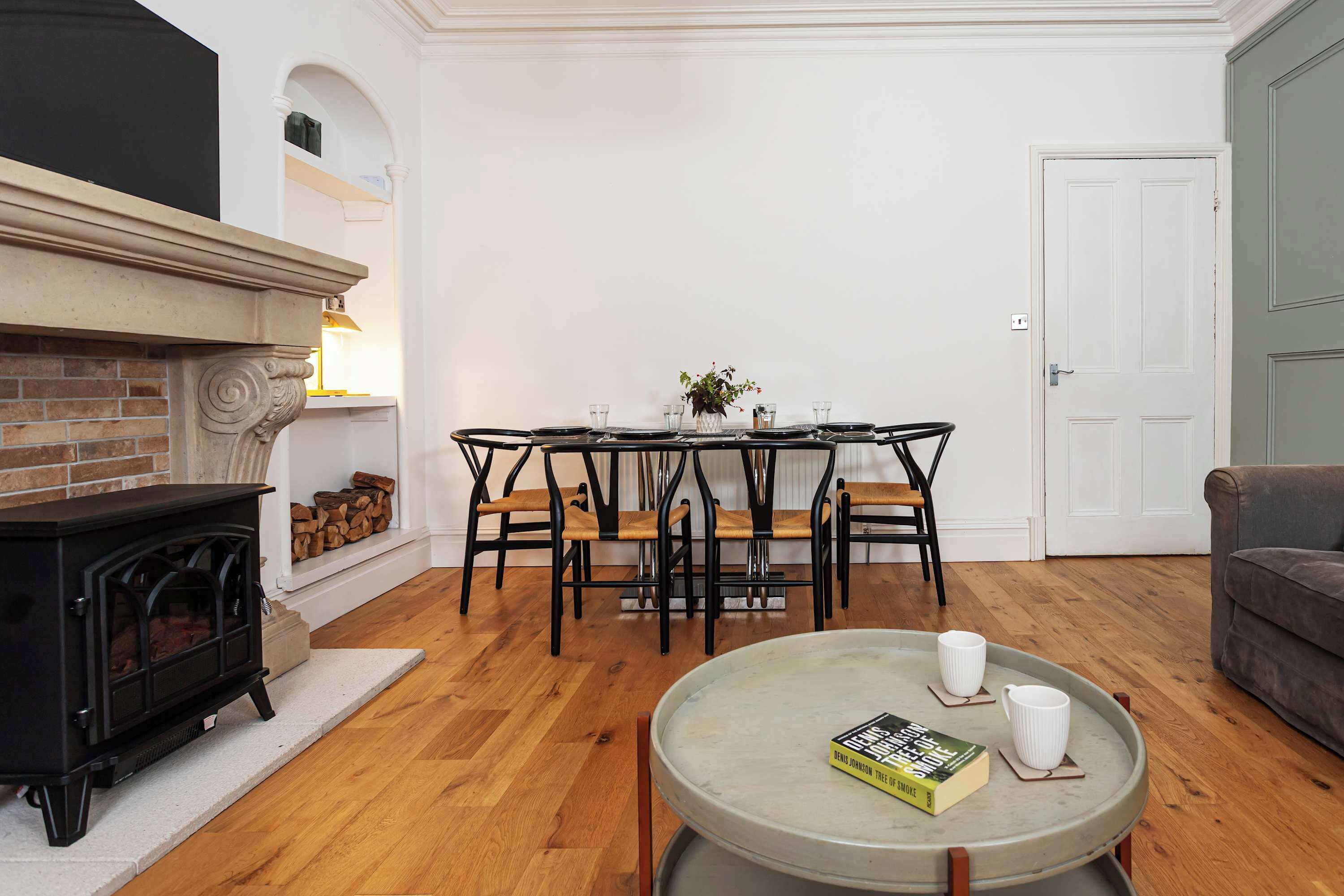 A living room with a fireplace, a dining table with chairs, a coffee table with a book and a mug, and a door.