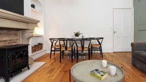 A living room with a fireplace, a dining table with chairs, a coffee table with a book and a mug, and a door. - Charming 2BR Flat on Grange-Over-Sands High Street (Grange over Sands)
