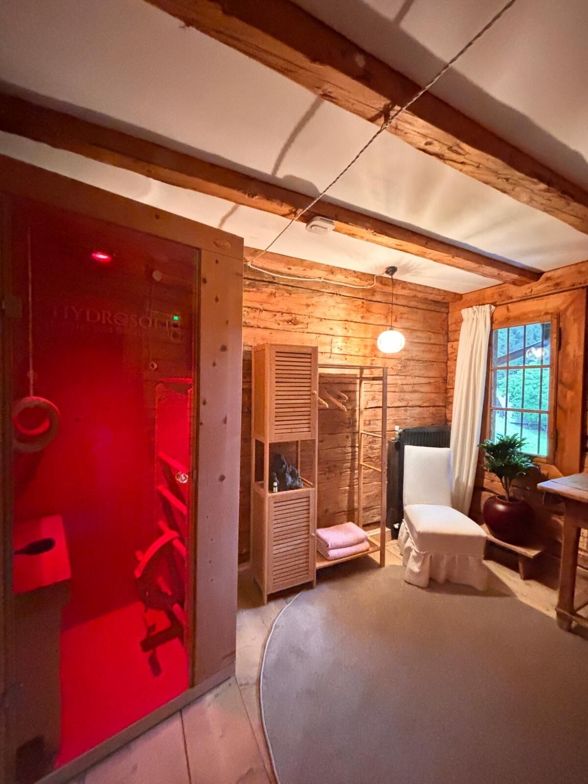 Una habitación con paredes de madera, una puerta iluminada con luz roja, una silla blanca, una mesa y una ventana con vistas.