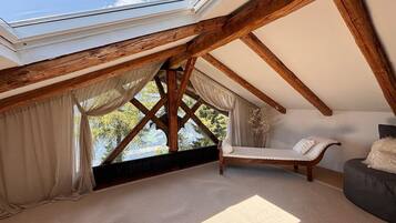 Ein Zimmer mit schrägen Wänden, Holzbalken, einem Oberlicht, einer gepolsterten Bank, einem Sofa und Blick ins Grüne.