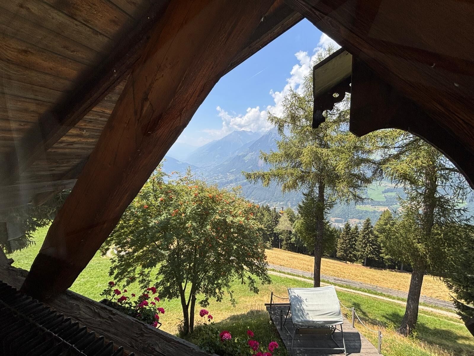 A wooden structure with a view of a mountainous landscape, a bench, and flowering plants.