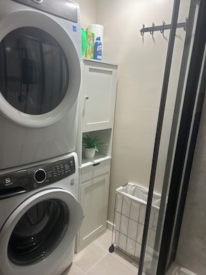 A laundry area with stacked washers and dryers, a small cabinet with a plant, and a metal laundry basket.
