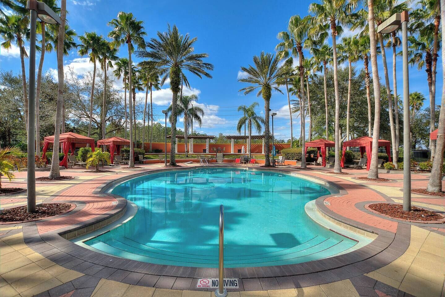 A swimming pool with a no diving sign, surrounded by palm trees and red canopied seating areas.