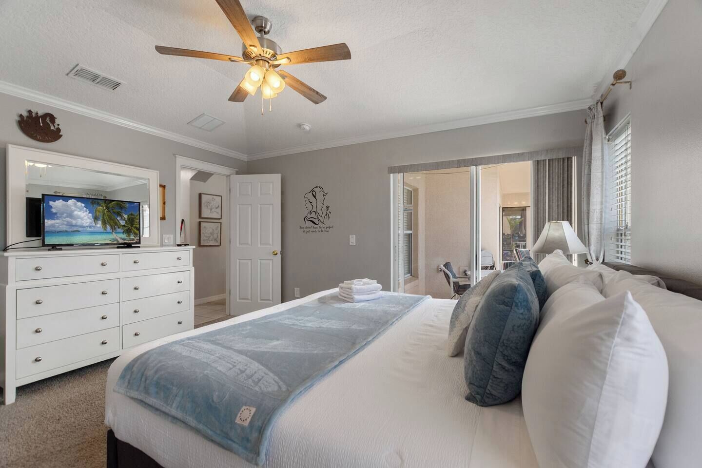 A bedroom with a bed, a television on a dresser, a ceiling fan, and a view of the outside.