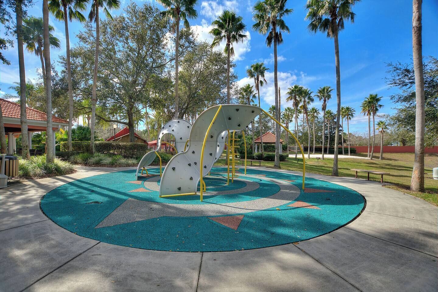 Ein Spielplatz mit Klettergerüsten und einer runden, grüner Spielzone, umgeben von Palmen.