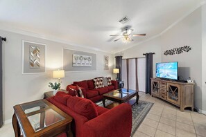 A living room with a red sofa set, a glass coffee table, a television on a wooden cabinet, and a ceiling fan. - Casa Sevilla | 4 BD | Sleeps 8 | Pool/Spa | Disney (Davenport)