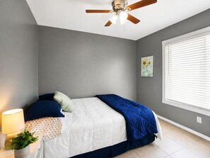 Een slaapkamer met een bed, een plafondventilator, een raam met jaloezieën en een wandlamp.