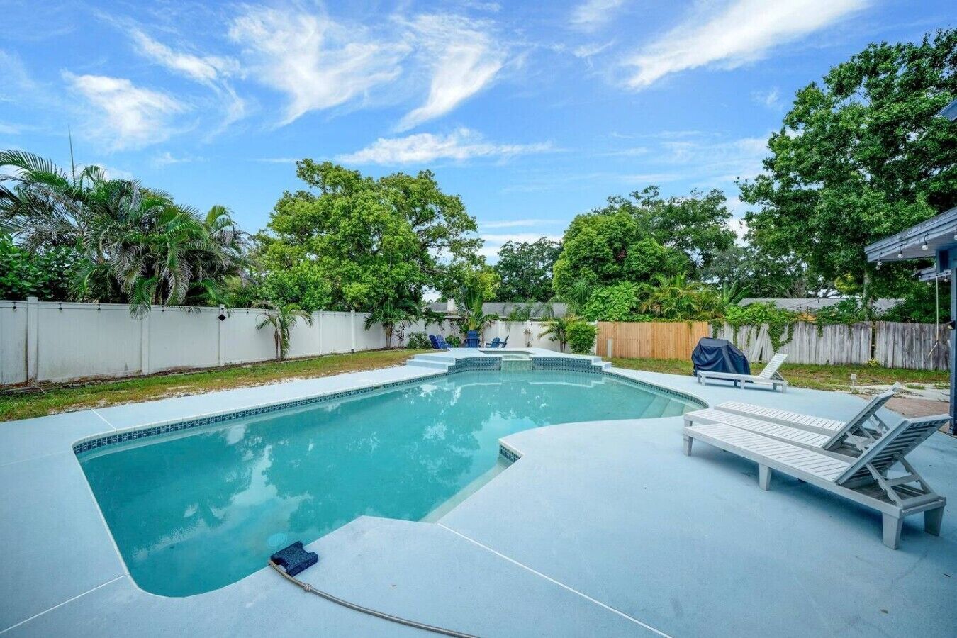 A rectangular swimming pool with a built-in hot tub, surrounded by a white fence and lush greenery.
