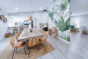 Un espace repas moderne avec une longue table, des chaises en osier et une fresque décorative sur le mur.