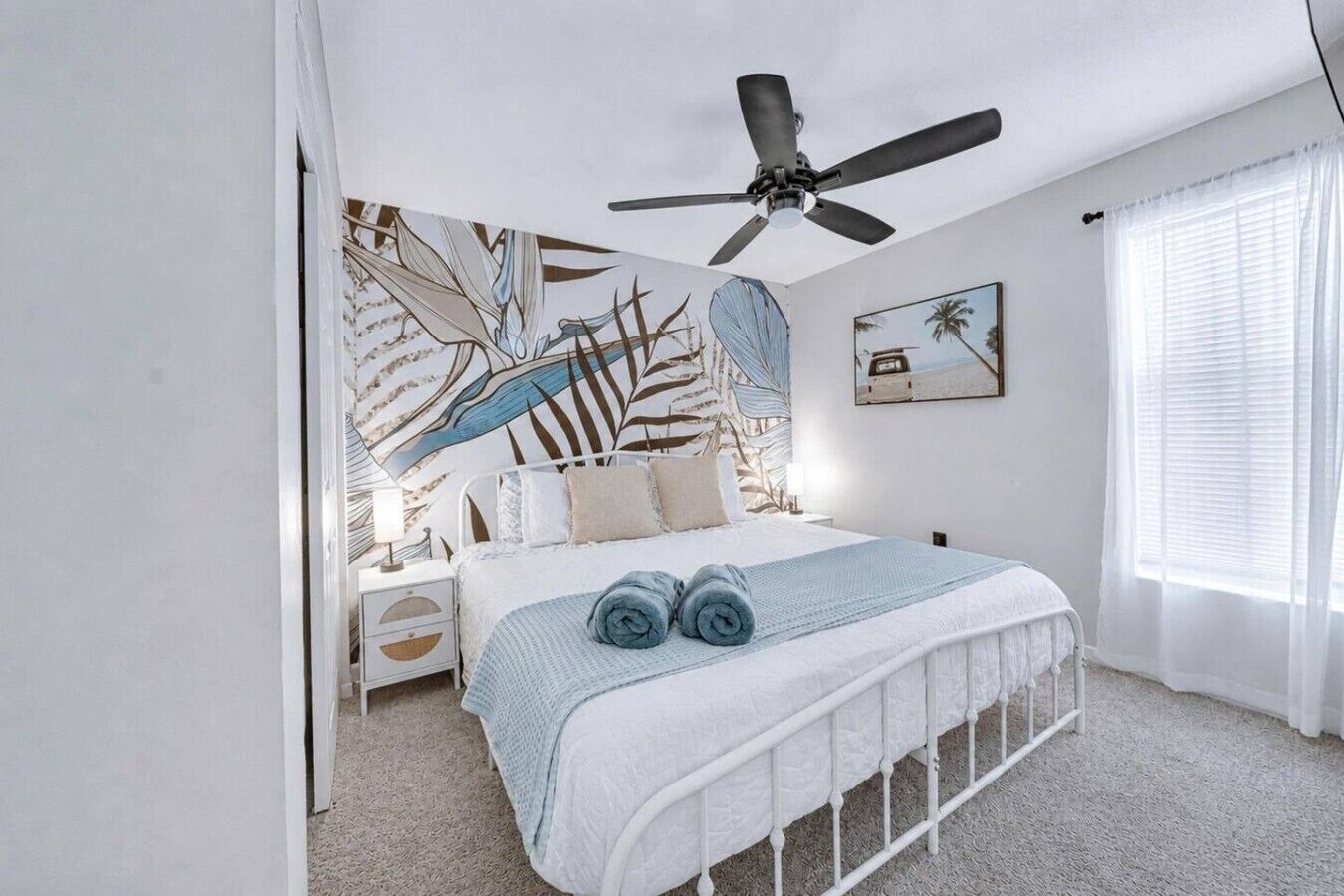 A bedroom with a bed, a ceiling fan, a nightstand, a wall with a tropical leaf pattern, and a window with blinds.