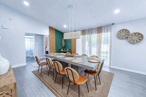 A modern dining area with a long table, multiple chairs, and a hanging light fixture. - Palm Harbor Pool 4BR | Hot Tub + BBQ + Game Room (Palm Harbor)
