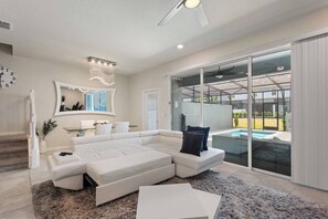 A modern living room with a sectional sofa, a large mirror, and a view of a swimming pool through glass doors. - Solterra 5BD•Sleeps13•Private Pool•Disney•Resort (Davenport)