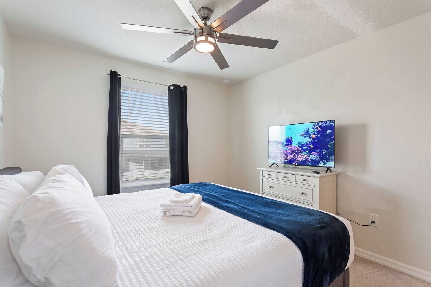 Habitación de hotel con cama, televisión, ventilador de techo y ventana con persianas.