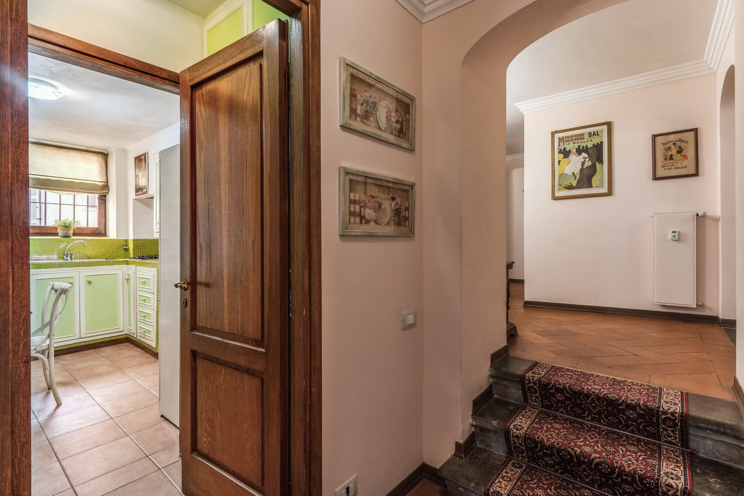 Un couloir avec une porte en bois, un escalier recouvert d’une moquette à motifs et une cuisine visible à travers une porte ouverte.