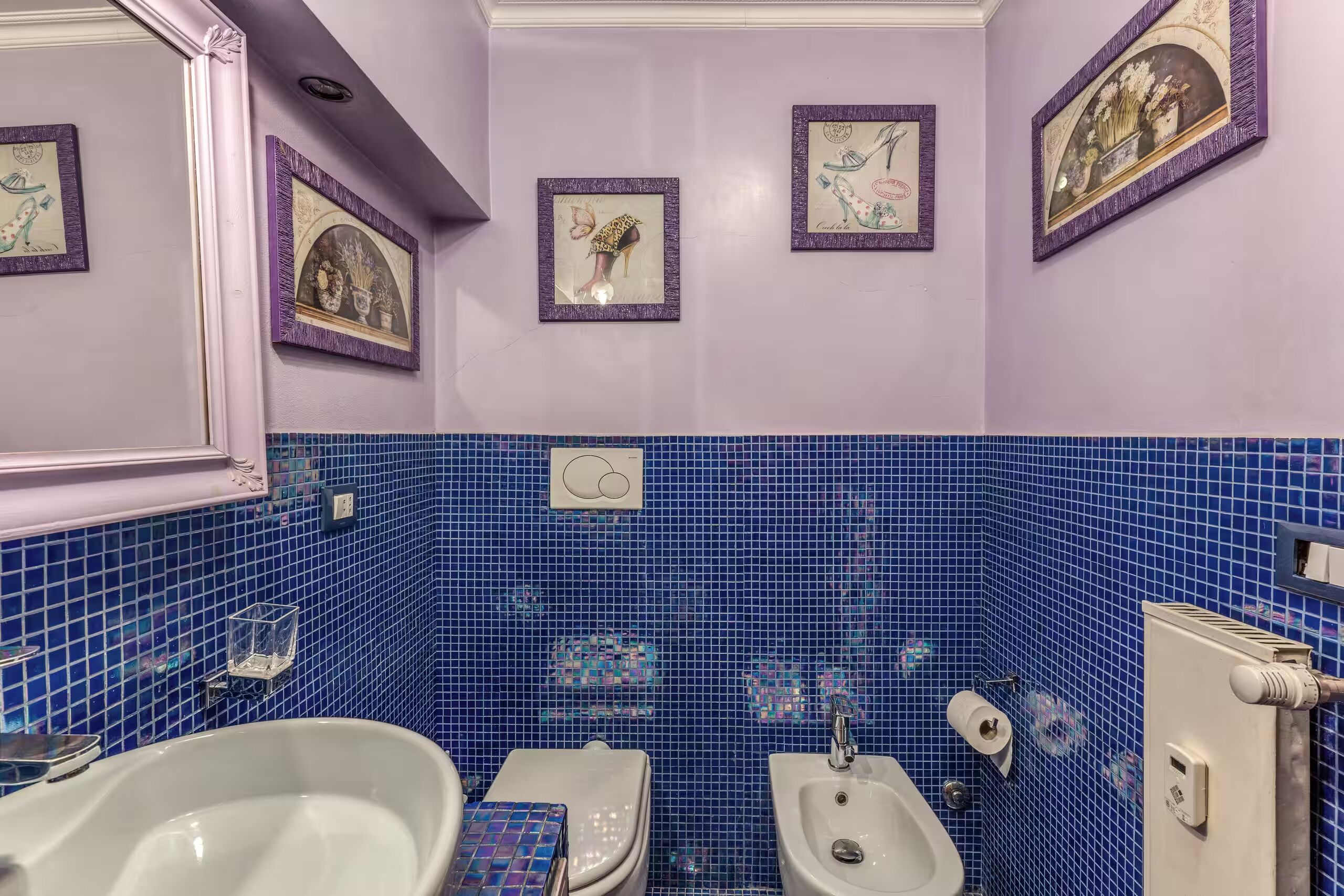 Un baño con pared de mosaicos azules, un lavabo blanco, un bidé y un inodoro. Hay cuadros enmarcados en la pared.