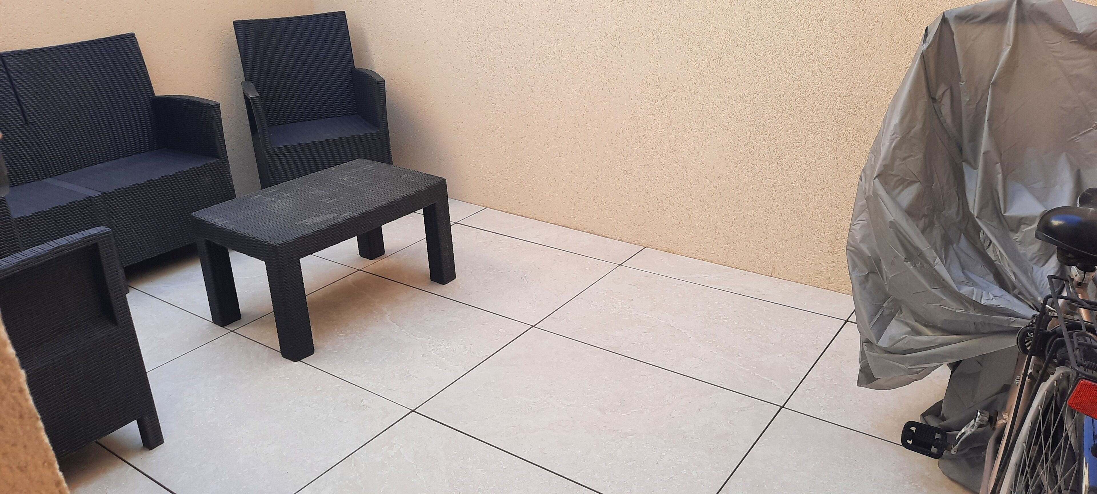 Uma pequena área de pátio com duas cadeiras de vime, uma mesa, uma bicicleta coberta e piso de azulejos.