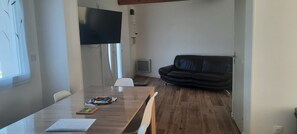 A room with a wooden table, a black leather sofa, a white chair, and a flat-screen TV mounted on the wall.
