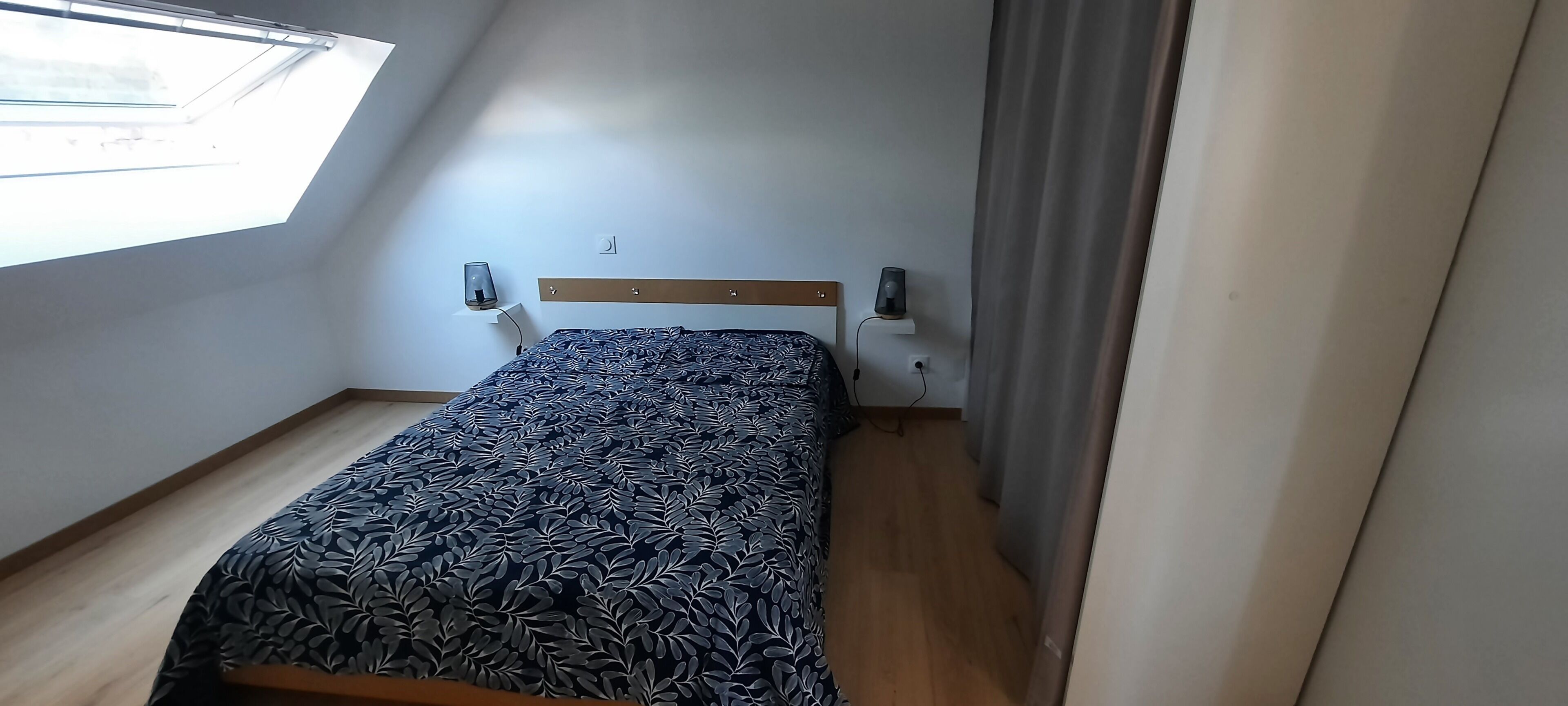 Pequeña habitación con cama individual, techo inclinado, ventana, mesita de noche, lámpara y toma de corriente.