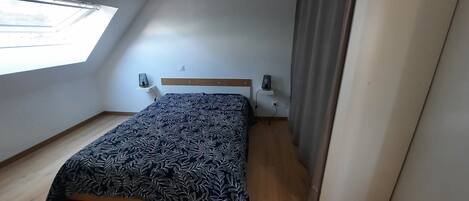 Une petite chambre avec un lit simple, un plafond en pente, une fenêtre, une table de chevet, une lampe et une prise de courant.
