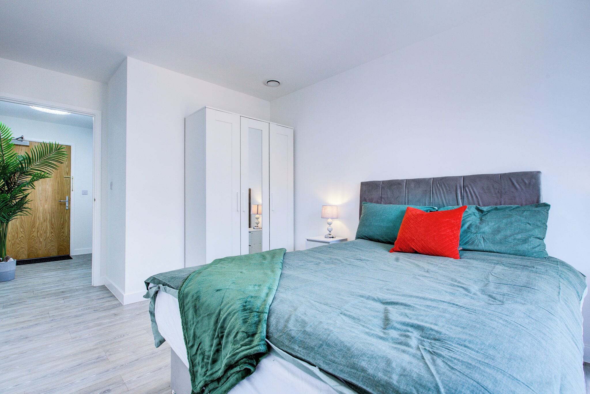 A modern bedroom with a bed, a wardrobe, a bedside table, a lamp, and a potted plant.