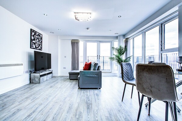 A modern living room with a flat-screen TV, grey sofa, dining table, and chairs.