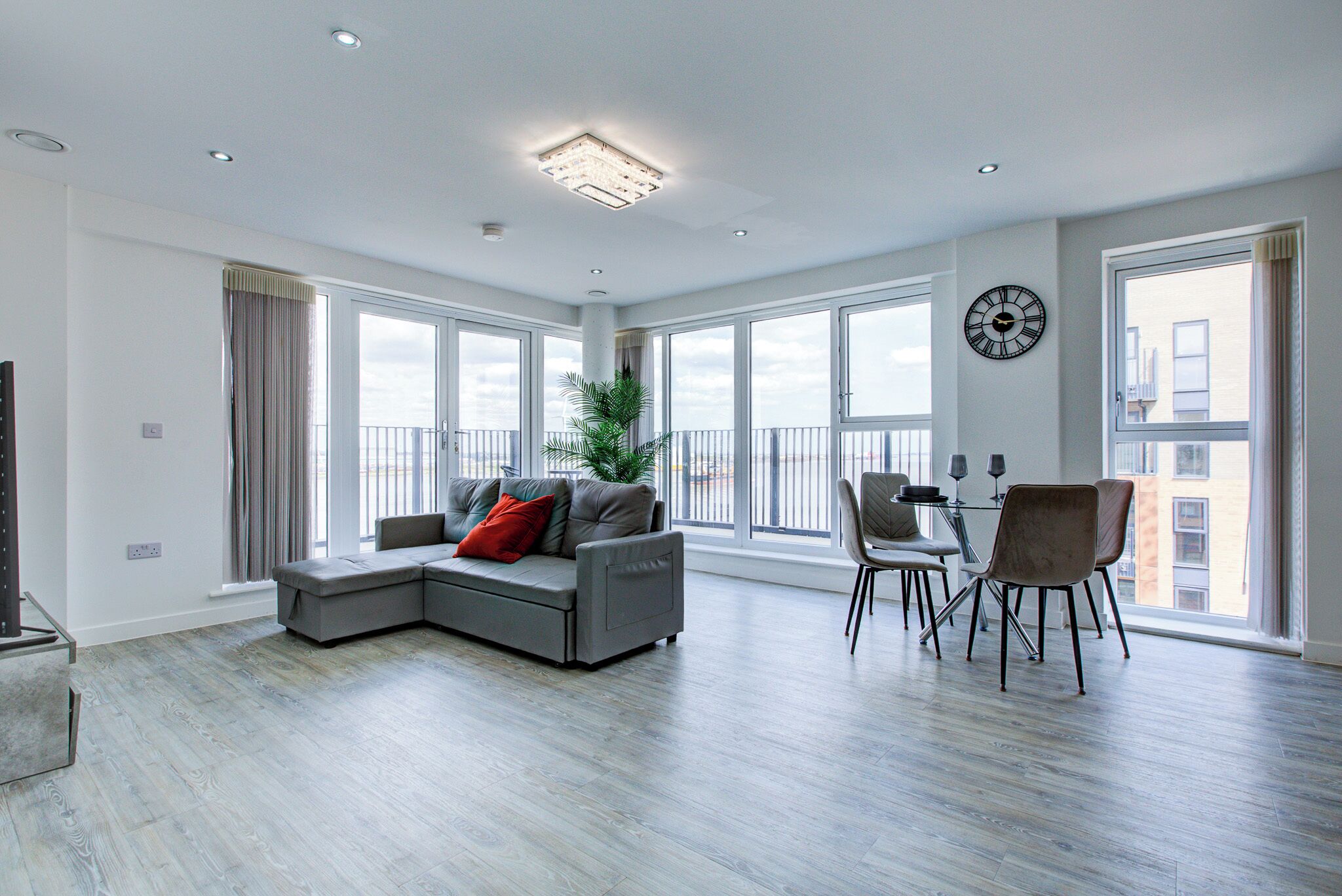 A modern living room with a sectional sofa, dining table, chairs, large windows, and a clock on the wall.
