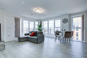 En moderne stue med en hjørnesofa, spisebord, stole, store vinduer og et ur på væggen.