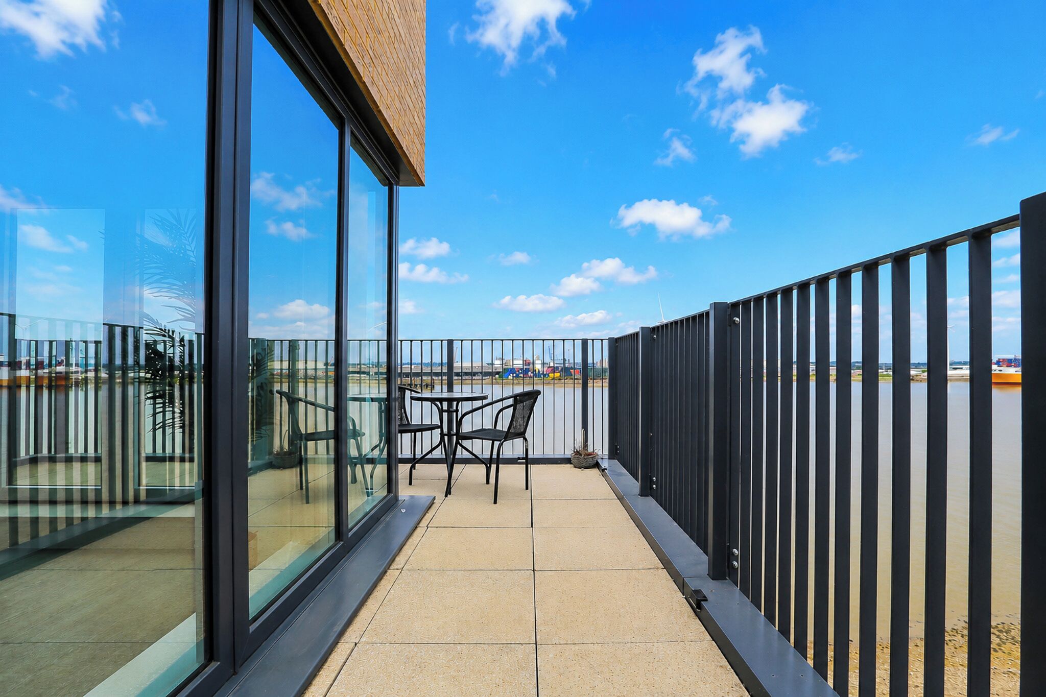 A balcony with a table and chairs, a glass wall, and a view of the sky and distant structures.