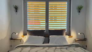 Une chambre à coucher avec un lit, deux tables de chevet, une fenêtre avec des stores et des plantes fixées au mur.