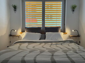 Ein Schlafzimmer mit einem Bett, zwei Nachttischen, einem Fenster mit Jalousien und Pflanzen an der Wand.