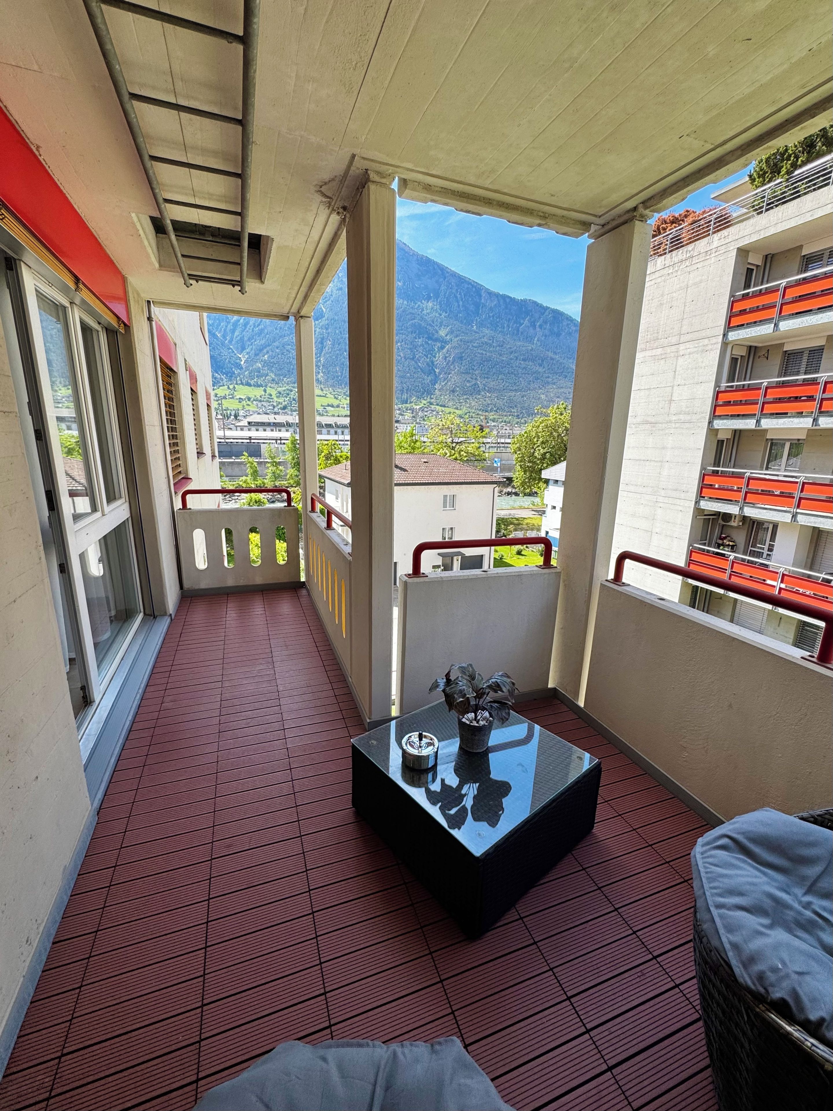 Un balcon avec un sol rouge, une table en verre ornée d’une plante, et une vue sur une montagne et des bâtiments.