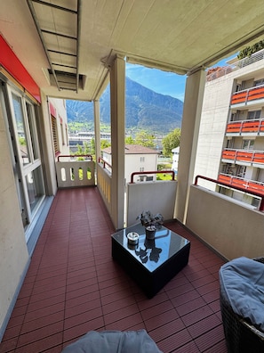 Ein Balkon mit rotem Boden, einem Glastisch mit Pflanze und Blick auf einen Berg sowie Gebäude.