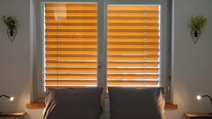 Une chambre à coucher avec un lit, deux tables de chevet avec des lampes, une fenêtre avec des stores et une petite plante sur chaque mur.